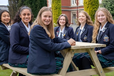 Un grupo de alumnas de la Otago Girls High School está sentado en un banco de madera en el área del jardín con árboles y edificios escolares al fondo.