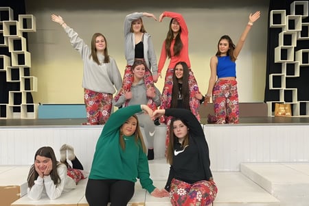 Un grupo de jóvenes estudiantes del Our Lady Catholic College está en trajes coloridos sobre un escenario con un fondo geométrico.