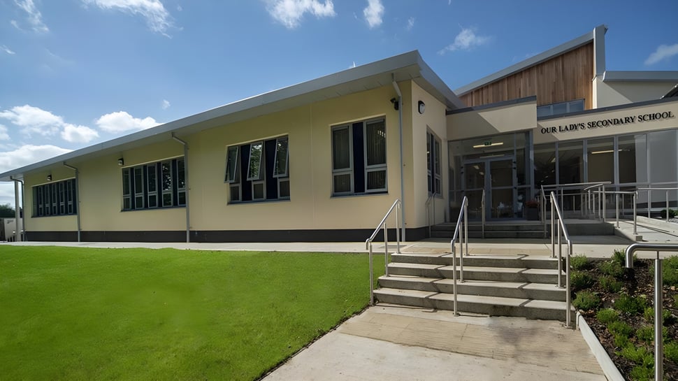 El moderno edificio escolar de Our Lady’s Secondary School con césped y escaleras frente a la entrada bajo un cielo azul.