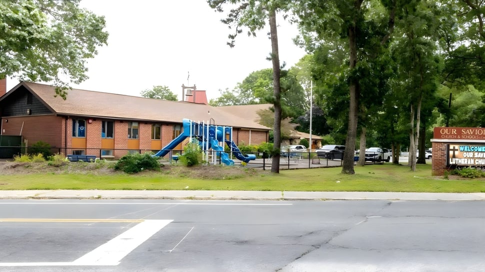 El edificio de madera de dos pisos de Our Savior New American School está rodeado de árboles verdes y se encuentra en una calle pavimentada.