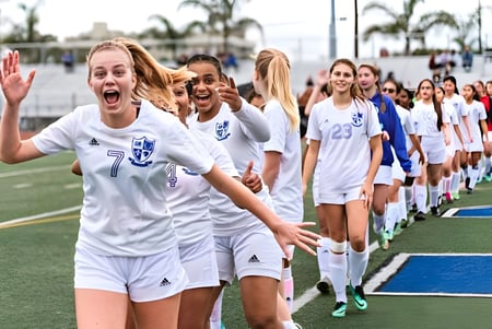 Un grupo de jugadoras de fútbol femenino está en el campo de deportes de la Pacifica Christian High School frente a palmeras y edificios.