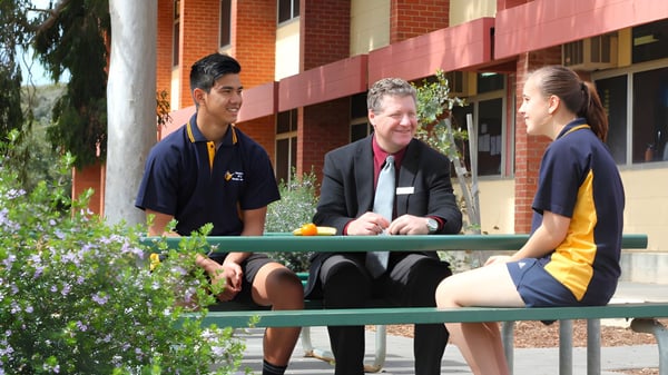 Dos estudiantes están sentados con una persona adulta en un banco frente a un edificio de ladrillo de la Paralowie R-12 School.
