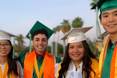 Un grupo de estudiantes de la Phoenix Christian Preparatory School está de pie en sus togas de graduación frente a un paisaje pintoresco con palmeras.