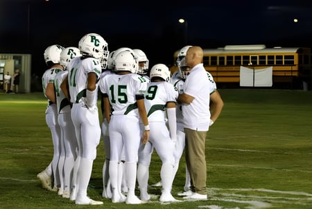 Estudiantes de la Phoenix Christian Preparatory School están juntos en camisetas blancas en un campo de fútbol débilmente iluminado con un autobús escolar al fondo.