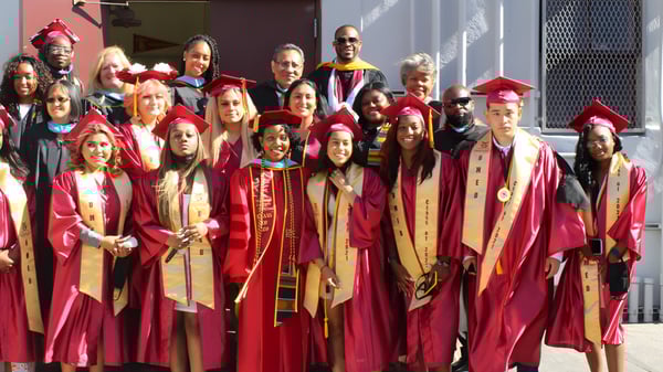 Un grupo de graduados de la Phoenix Magnet Academy está de pie en togas rojas y doradas frente a un edificio con fachada roja y blanca.