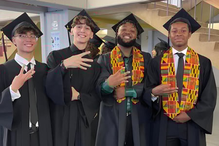 Cuatro estudiantes de la Pierrefonds Community High School en ropa de graduación posan juntos para una foto en el pasillo de la escuela.