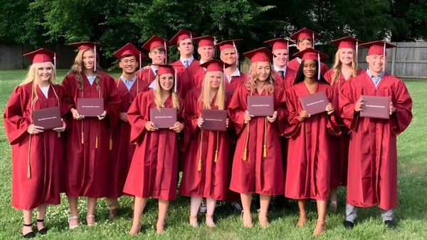 Un grupo de graduados de la Plaza Heights Christian Academy está al aire libre frente a árboles verdes en togas de graduación rojas.