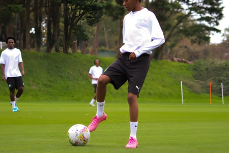 Un estudiante del Plymouth College dribla un balón de fútbol en un campo de césped con otros jugadores al fondo.