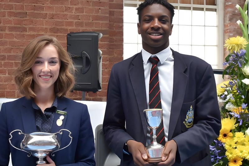 Dos personas en ropa formal sostienen trofeos en una ceremonia de premiación en el campus de la Pocklington School.