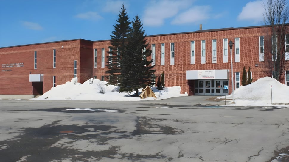 El gran edificio de ladrillo de la Polyvalente Alexandre-J.-Savoie se encuentra detrás de un alto pino en el invierno cubierto de nieve.