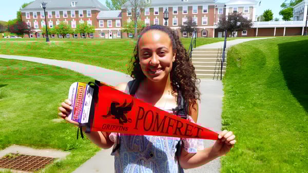 Una joven sonriente sostiene en la hierba frente a un edificio de ladrillo en el campus de la Pomfret School una bandera roja y blanca de Pomfret.