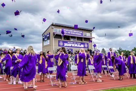 Los graduados de Portland Christian High School celebran con togas moradas y sombreros voladores en el campo deportivo.