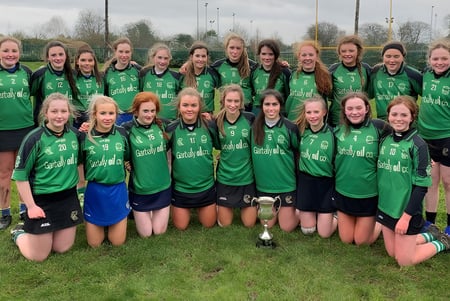 Un grupo de jugadoras de fútbol del Portumna Community College posan en un campo con un trofeo en primer plano.