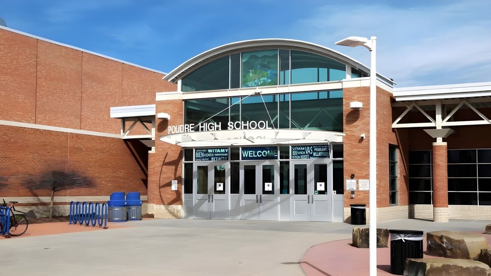El edificio de ladrillo con una entrada de vidrio arqueada en Poudre School District está rodeado de un área pavimentada con bancos y un soporte para bicicletas.