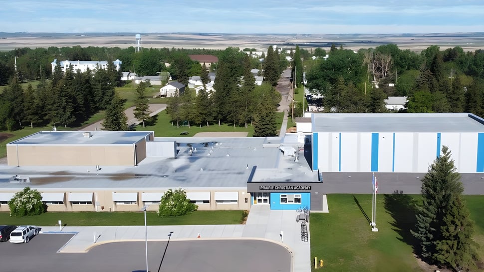 El moderno edificio principal de la Prairie Christian Academy se encuentra frente a un prado con un bosque al fondo bajo un cielo azul.