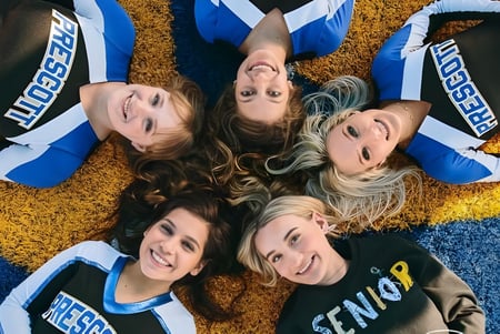 Jóvenes mujeres en uniformes de animadoras están sonriendo en el suelo frente a un fondo colorido en el campus de Prescott High School.