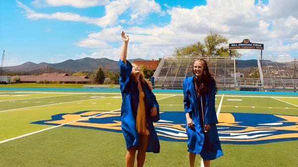 Dos graduados en togas de graduación azules están de pie en el campo deportivo de Prescott High School frente a montañas y un cielo nublado.