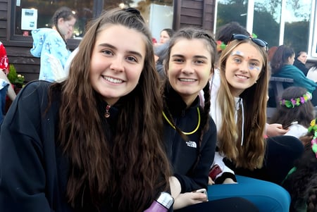 Tres estudiantes con cabello largo están sonriendo frente a un edificio con fachada de madera en el terreno de la Presentation Secondary School en Kilkenny.