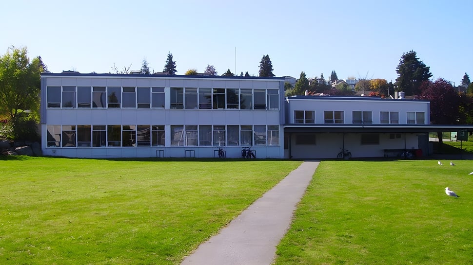 El moderno edificio de vidrio de la Prince of Wales Secondary School se encuentra en un campo cubierto de hierba bajo un cielo azul.