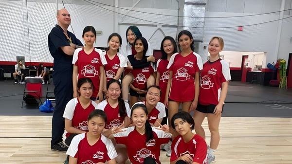Un grupo de jóvenes estudiantes en uniformes rojos posan en un gimnasio de la Princeton International School of Mathematics & Science con un hombre al fondo.