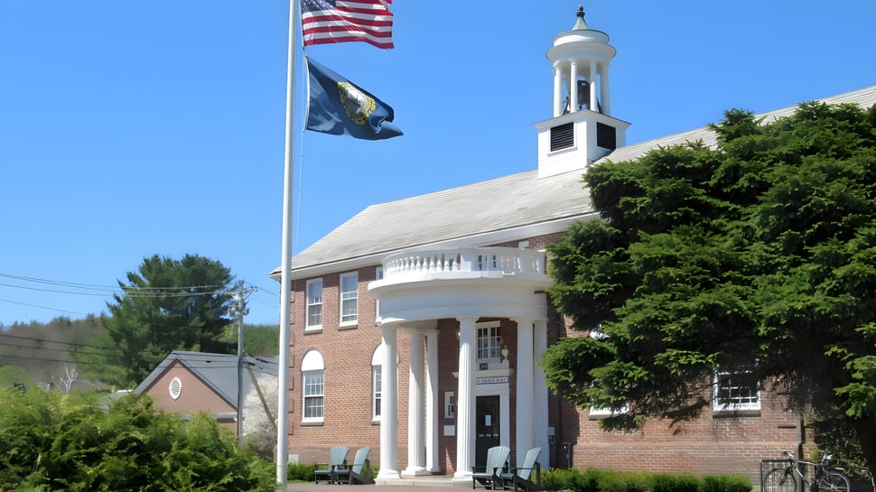 El edificio de ladrillo de la Proctor Academy con una torre blanca y dos banderas ondeando está rodeado de árboles.