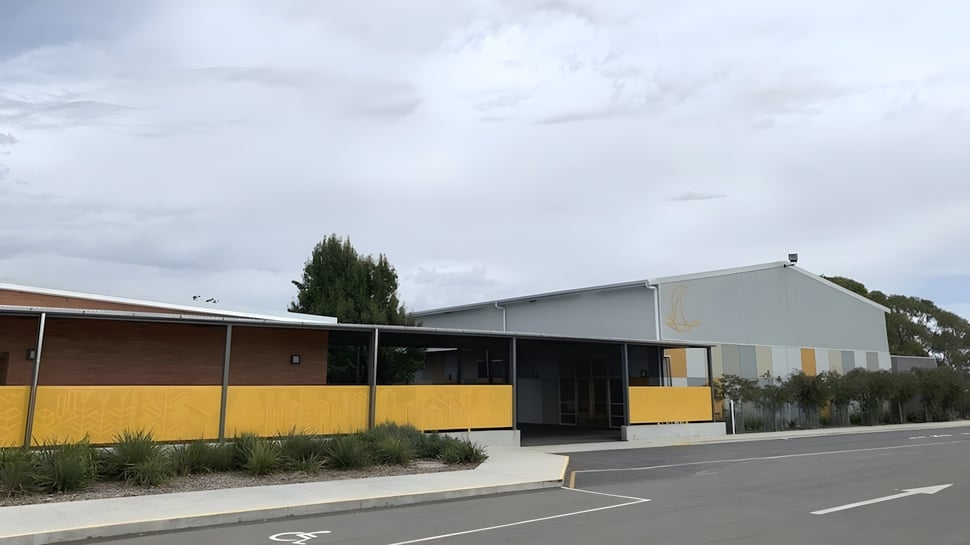 El edificio de una planta de la Prospect High School con fachada amarilla y entrada cubierta está rodeado de un estacionamiento y árboles.