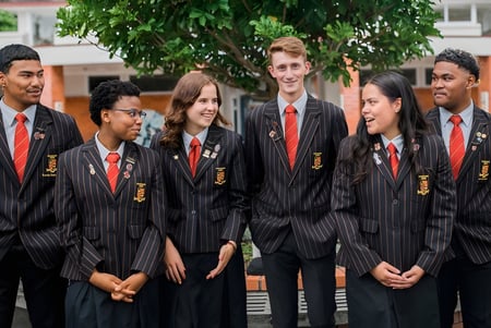 Un grupo de estudiantes en togas negras y corbatas rojas está frente a árboles verdes en el terreno de la Pukehohe High School.