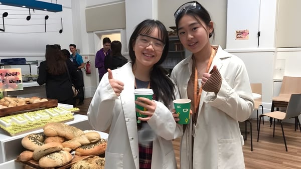 Dos estudiantes de The Purcell School for Young Musicians llevan batas de laboratorio y sostienen tazas de café en una habitación con pasteles.