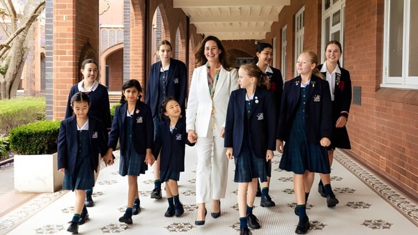 Un grupo de alumnas del Pymble Ladies College está en ropa formal juntas en un pasillo revestido de ladrillo con ventanas de arco.