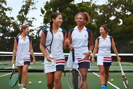 Un grupo de jóvenes tenistas está en el campo de tenis del Pymble Ladies College rodeado de árboles y áreas verdes.
