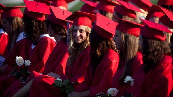 Un grupo de graduadas y graduados de la Quad Cities Christian School está en togas rojas con flores frente a un fondo verde.