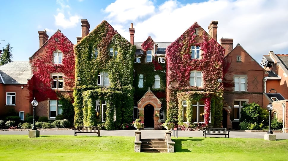El edificio de ladrillo histórico de la Queen Anne’s School está rodeado de hojas rojas y verdes y se encuentra detrás de un césped cuidado con bancos.