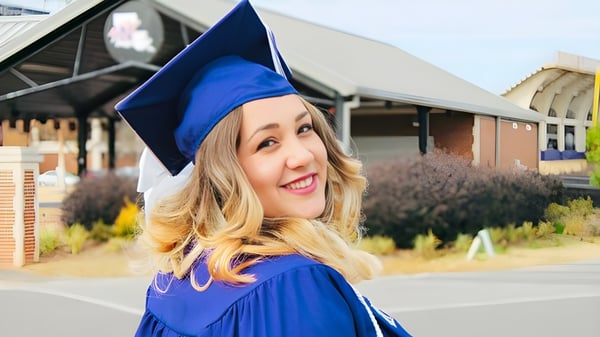 Una joven graduada de la Queen Elizabeth High School sonríe en su toga de graduación azul frente a un edificio del campus.