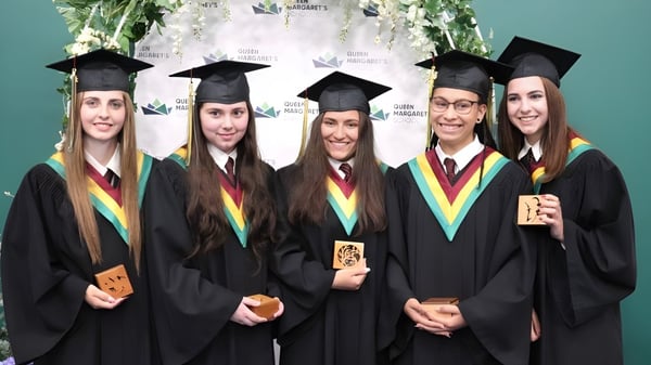 Cinco estudiantes de Queen Margaret’s School están en sus togas de graduación y sostienen sus diplomas frente a un logo universitario.