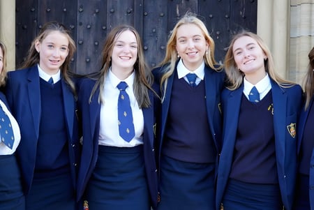 Seis alumnas en uniformes escolares sonríen frente a una puerta de madera en el campus de la Queen Mary's School.