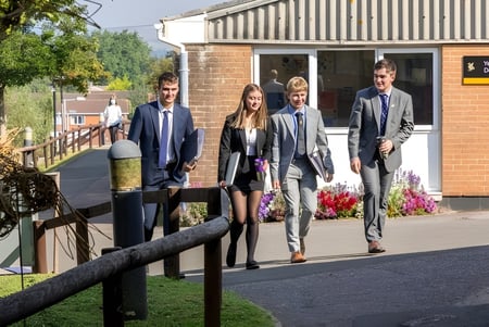 Un grupo de personas camina a lo largo de un camino en el campus de Queen’s College con un entorno verde y edificios de fondo.