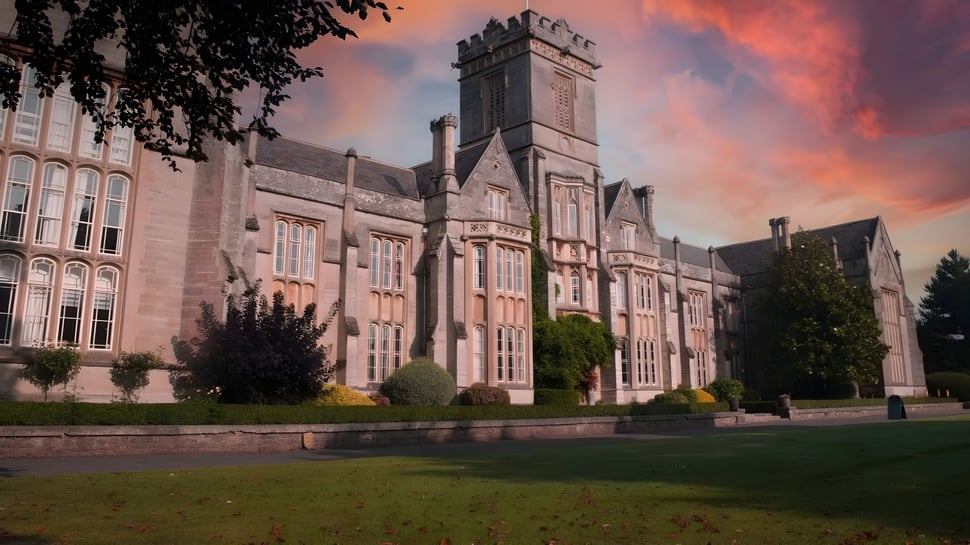 El magnífico edificio principal de Queen’s College está rodeado de naturaleza verde y se encuentra frente a un colorido cielo de atardecer.
