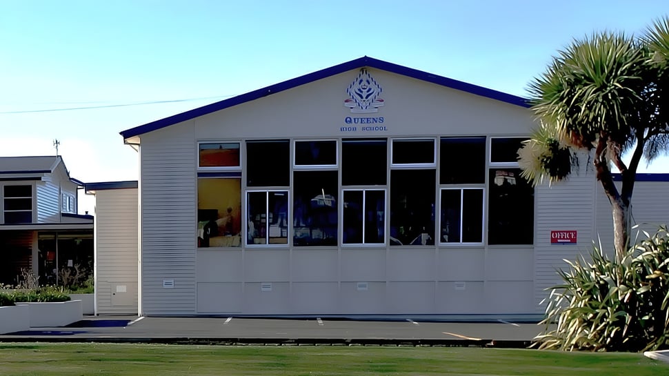 Un edificio de madera con techo azul se encuentra frente a palmeras en el terreno de la Queens High School.
