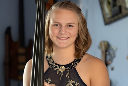 Una estudiante de la Queensbury High School sostiene un cello en una sala con obras de arte en las paredes.