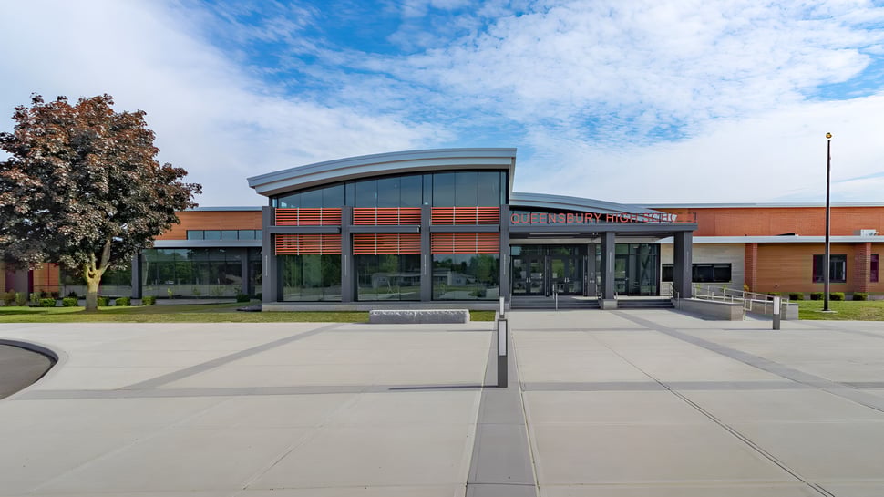 El moderno edificio con fachada de vidrio de la Queensbury High School se encuentra en un patio pavimentado rodeado de árboles.
