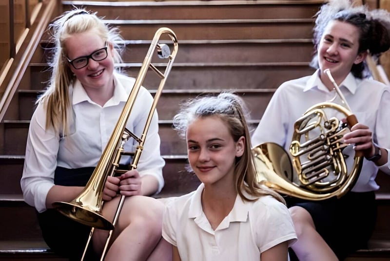Tres estudiantes de la Queenswood School están con un trombón y un cuerno francés en una escalera de madera frente a un fondo de madera.