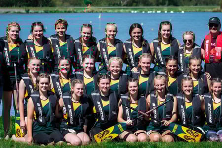 Un grupo de jóvenes deportistas en camisetas verdes y amarillas posan frente a un lago con árboles en el terreno de la Rangiora High School.