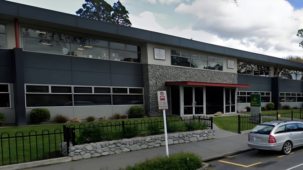 El moderno edificio de varios pisos de la Rangiora High School con fachada de piedra está rodeado de áreas verdes y un auto estacionado.