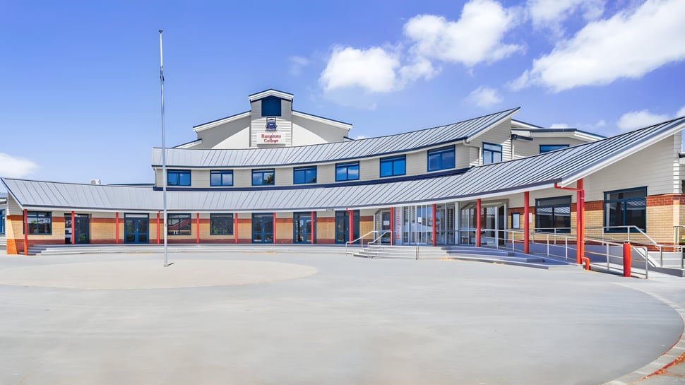 El moderno edificio de varios pisos con techo curvado y grandes ventanas del Rangitoto College está rodeado de un área exterior pavimentada.