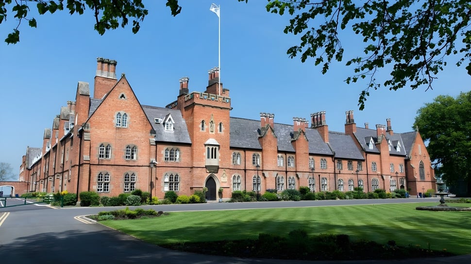 El gran edificio de ladrillo con torres y chimeneas en el terreno del Ratcliffe College está rodeado de césped bien cuidado y árboles.