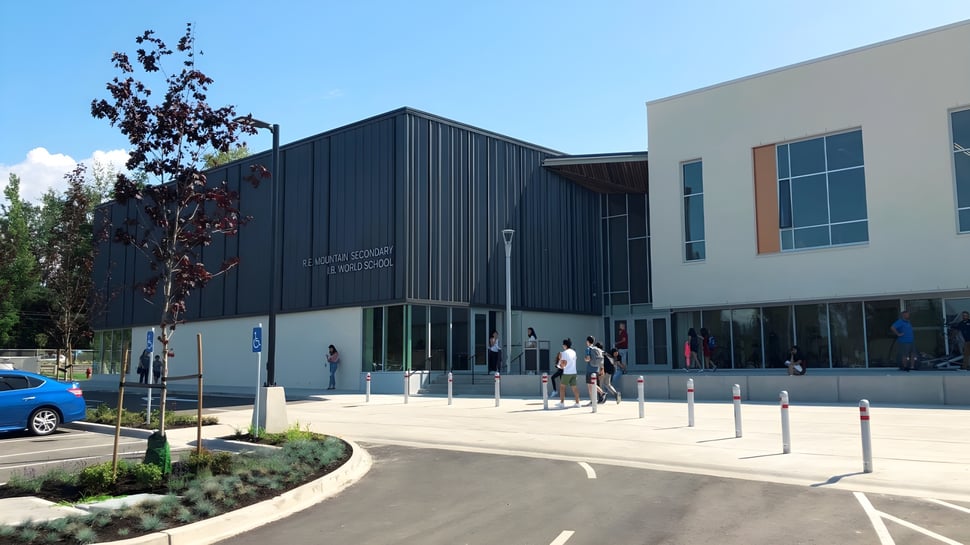 Se puede ver el moderno edificio de varios pisos de la R.E. Mountain Secondary School con fachada oscura y área pavimentada circundante.