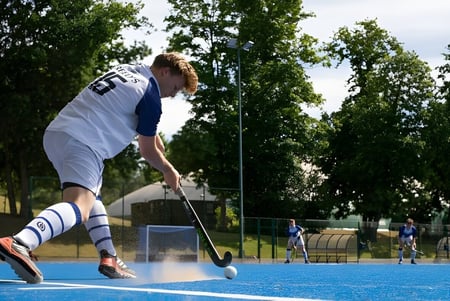 Un estudiante de la Reed’s School juega al hockey sobre césped en un campo de césped artificial azul con otros jugadores al fondo.