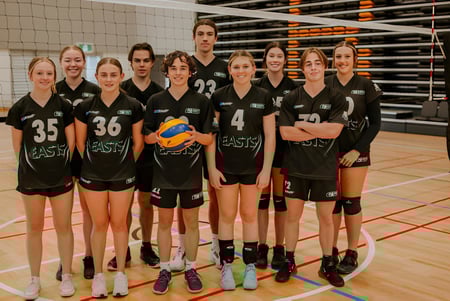 Un grupo de jóvenes jugadoras de voleibol del Reynella East College está reunido en la cancha de voleibol con la red y el marcador al fondo.