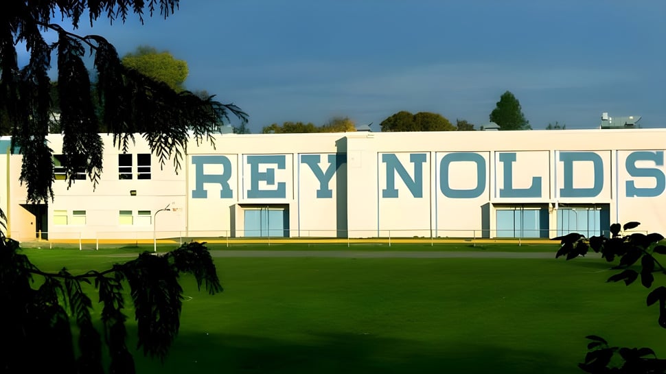 El gran edificio blanco de la Reynolds Secondary School está rodeado de vegetación verde y un campo de césped.