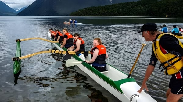 Estudiantes de la Riccarton High School están sentados con chalecos salvavidas en un bote en un lago entre montañas y bosques.